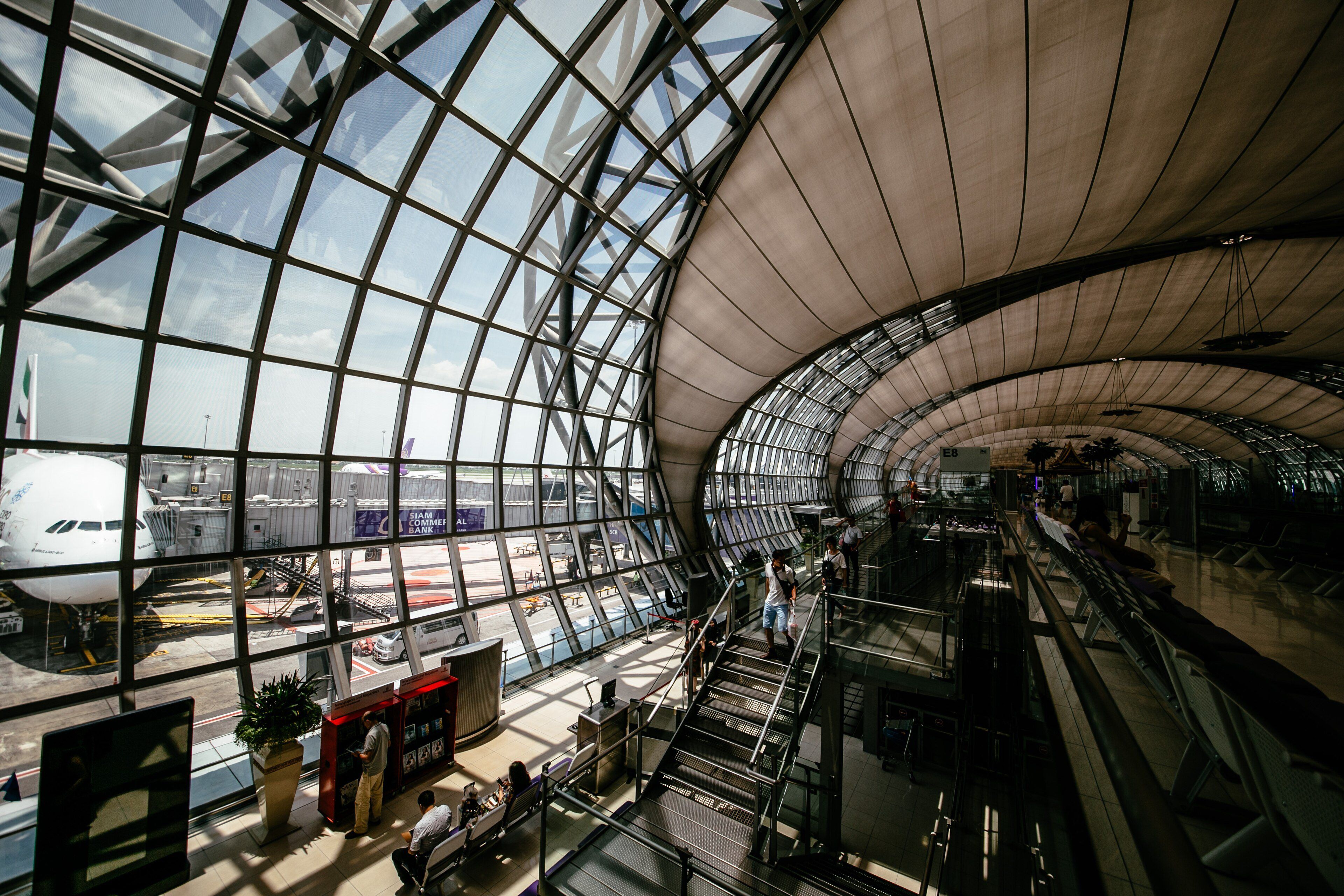 A glass and steel airport terminal hall with travelers and a parked aircraft outside