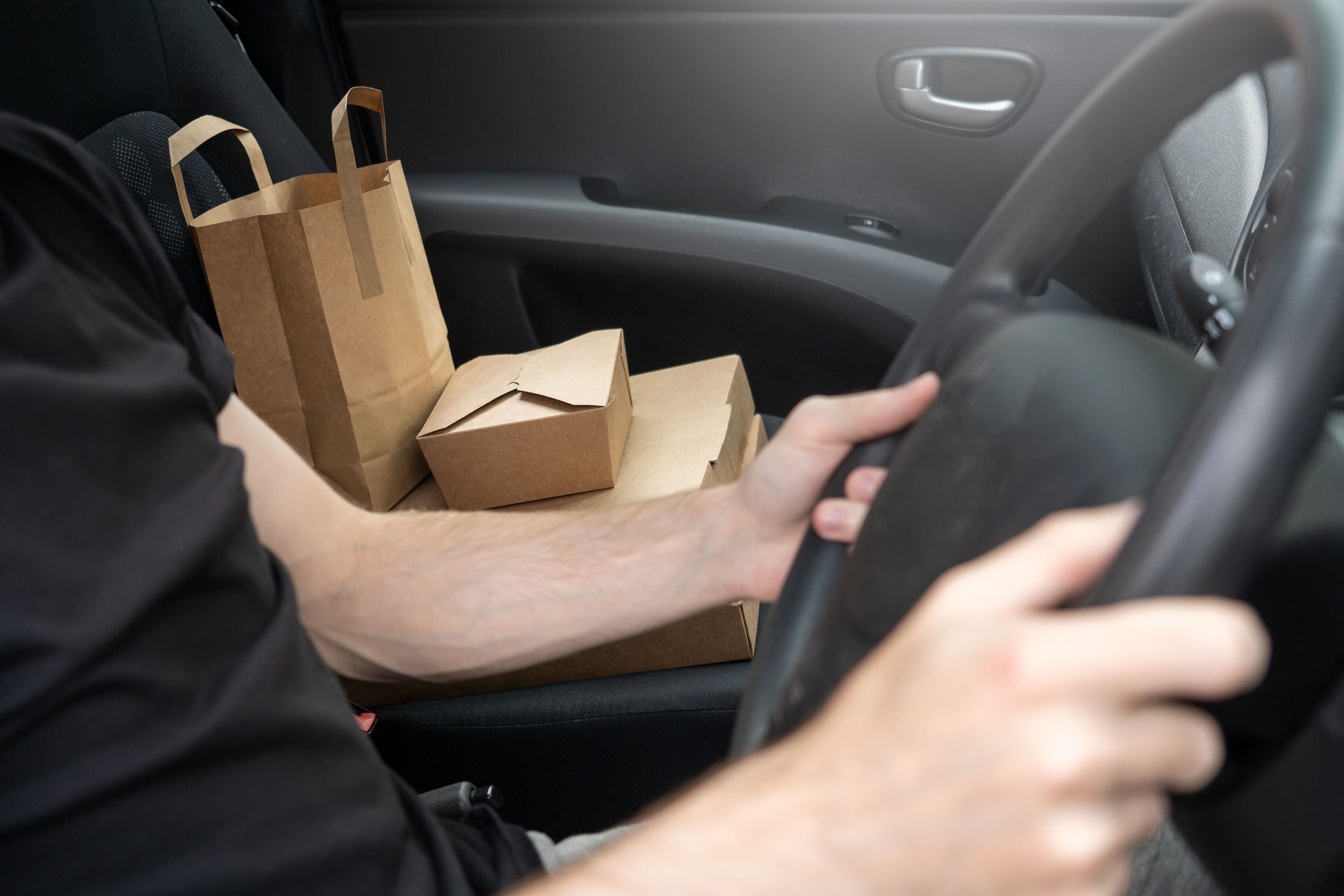 Takeout boxes and a paper bag on a car's passenger seat while the driver holds the wheel