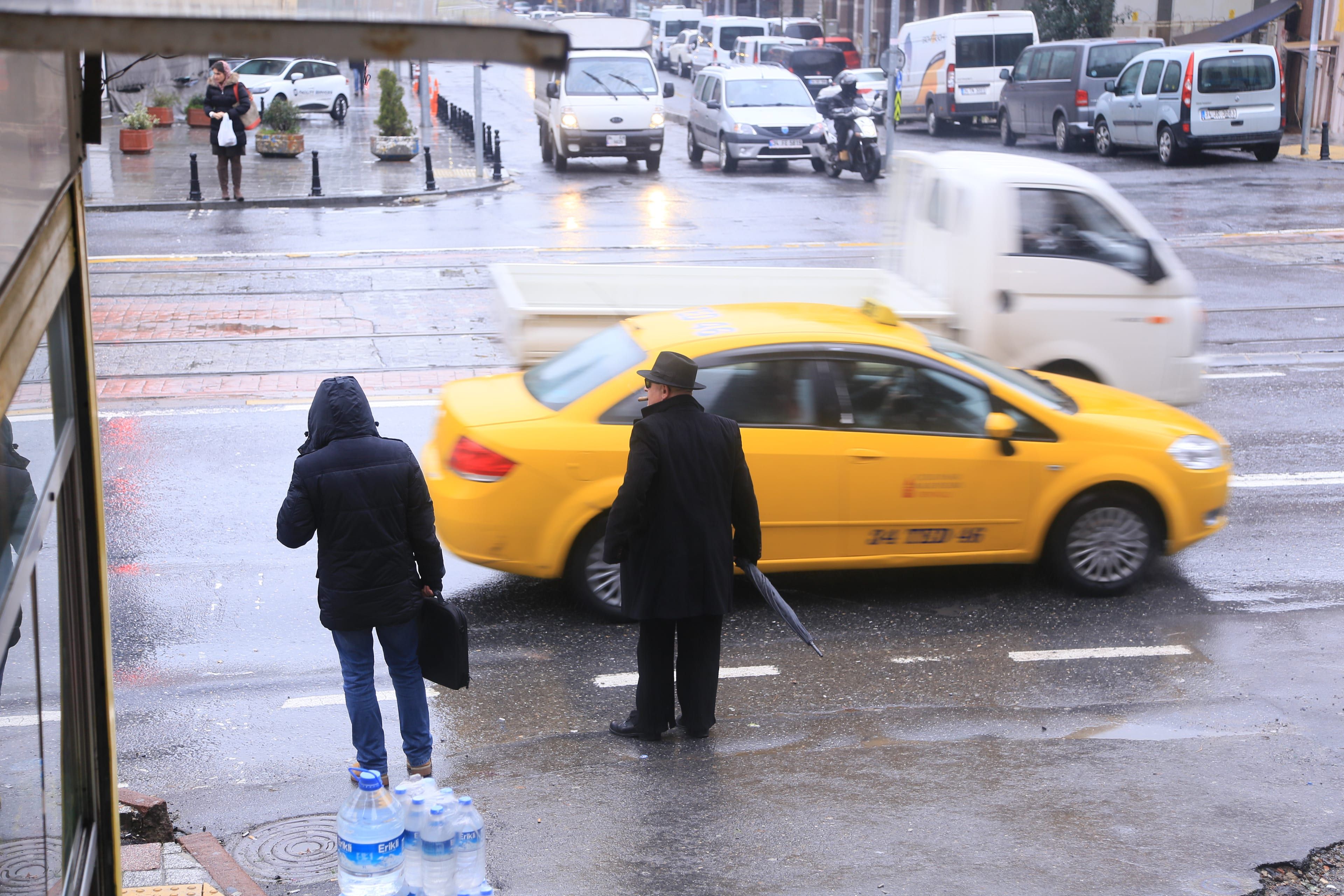 A yellow taxi turns on a wet street in Istanbul while two people wait under the rain